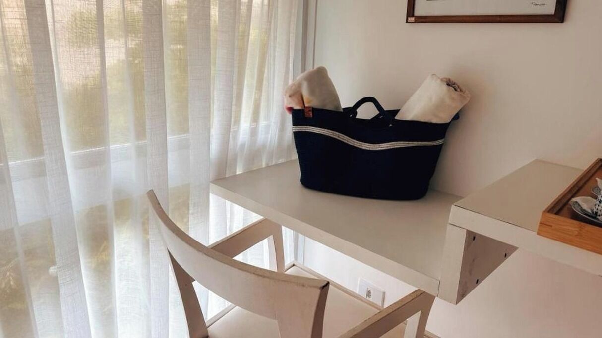 A room with a white desk, a dark blue bag on the desk, and a window with sheer curtains.