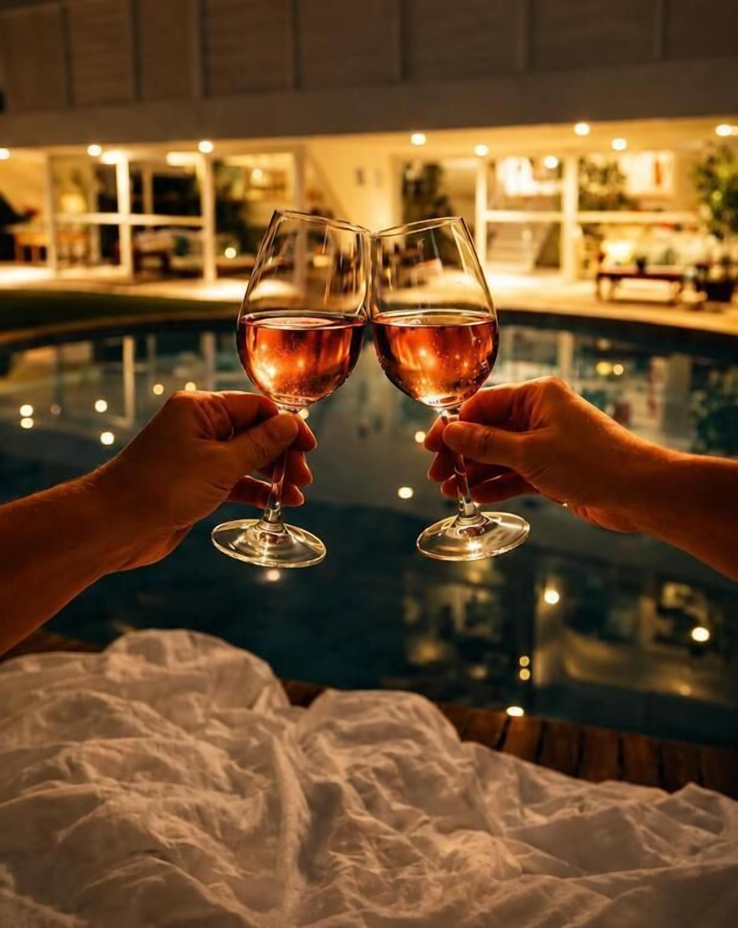 Two hands holding wine glasses over a pool with a building in the background.