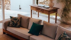 Un canapé beige avec trois coussins, une table d’appoint en bois avec une lampe, et une vue sur un mur en pierre et un espace extérieur.