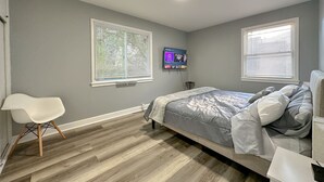 Une chambre à coucher avec un lit, une chaise, un téléviseur et deux fenêtres équipées de stores.