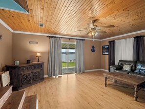 Ein Zimmer mit Holzboden, Deckenventilator, Holztisch, Ledersofa, Holzschrank und Seeblick durch eine Glastür.