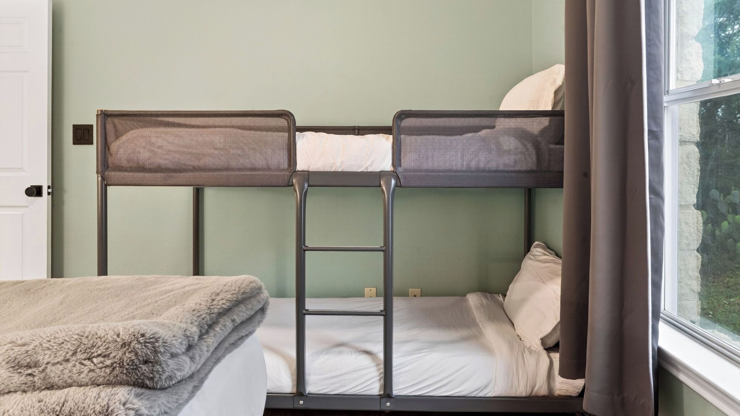 A bunk bed with a ladder, a single bed with a blanket, and a window with a view of greenery.