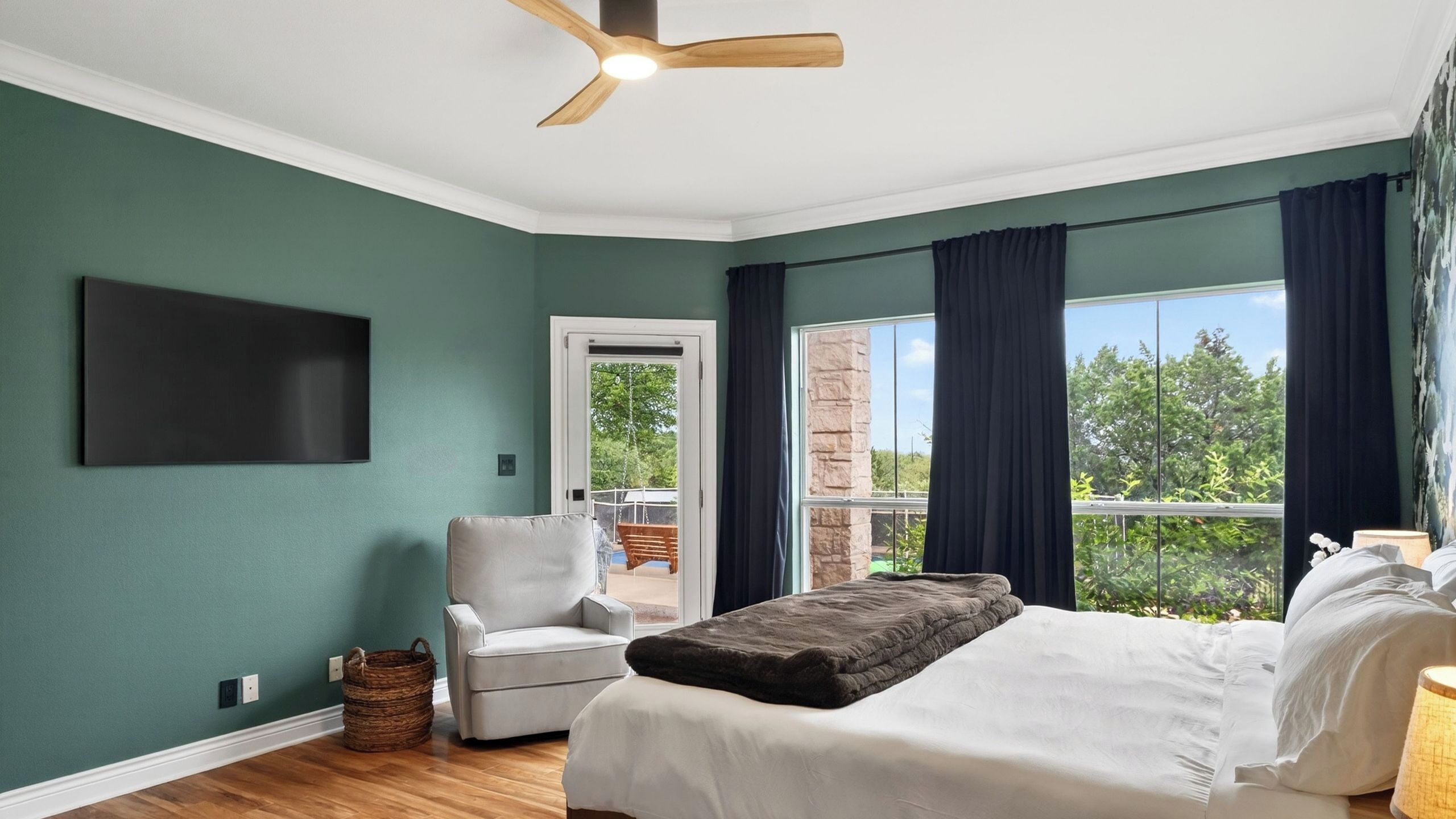 A bedroom with a bed, a chair, a ceiling fan, a TV, and a view of the outdoors.