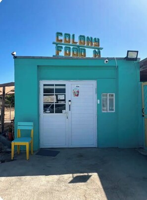 A small food stand with a sign that reads 'Colony Food M.' There is a white door with a window, a small chair, and a security camera on the roof.
