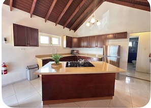 A kitchen with a central island, wooden cabinets, a stainless steel refrigerator, and a ceiling with exposed beams.