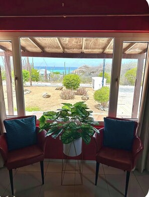 A room with a view of a garden and a beach. There are two chairs with cushions and a potted plant in the center.