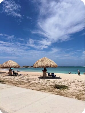Une scène de plage avec deux parasols en chaume, des chaises longues et un ciel bleu dégagé.