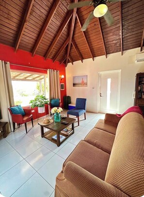 Un salon avec un plafond haut, un canapé, une table basse et un fauteuil. Il y a une porte et une fenêtre avec des rideaux.