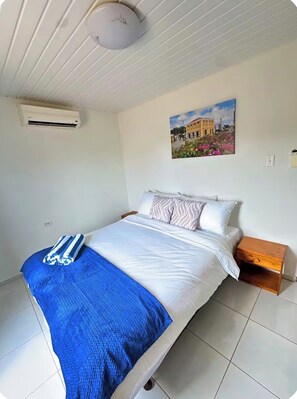 A hotel room with a bed, a nightstand, an air conditioning unit, and a wall-mounted picture.