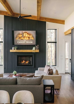 A modern living room with a fireplace, a ceiling fan, and a large window. - The Canopy House - Hot Tub, Sauna, Mountain View (Blue Ridge)