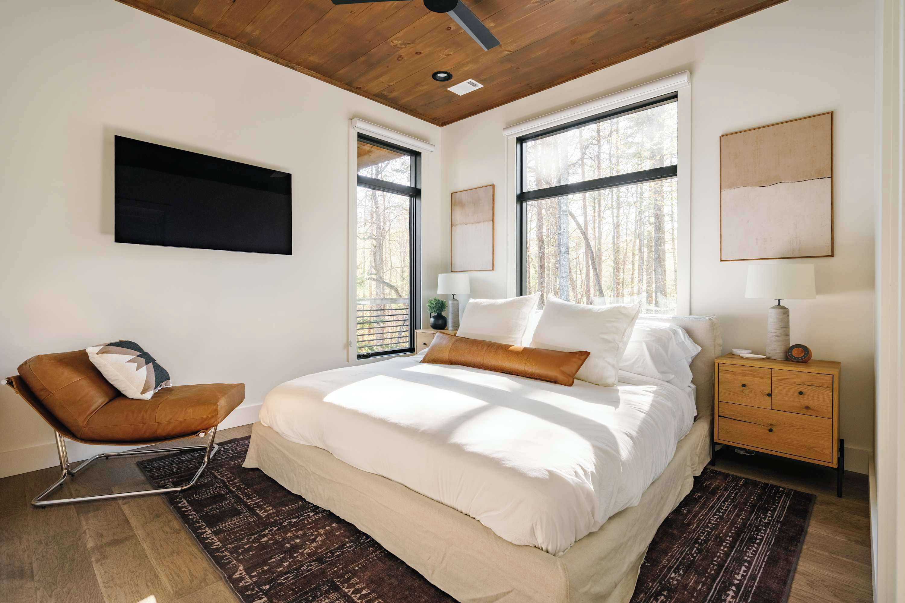 A bedroom with a bed, a chair, a nightstand, and large windows.