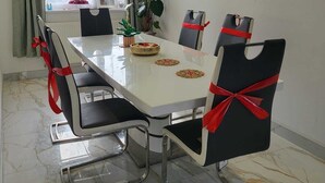 Comedor con una mesa blanca, sillas negras con lazos rojos, una planta en maceta y un ventanal con cortinas.