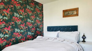 Un dormitorio con una cama, cabecera, mesita de noche, lámpara y un papel tapiz floral en la pared.