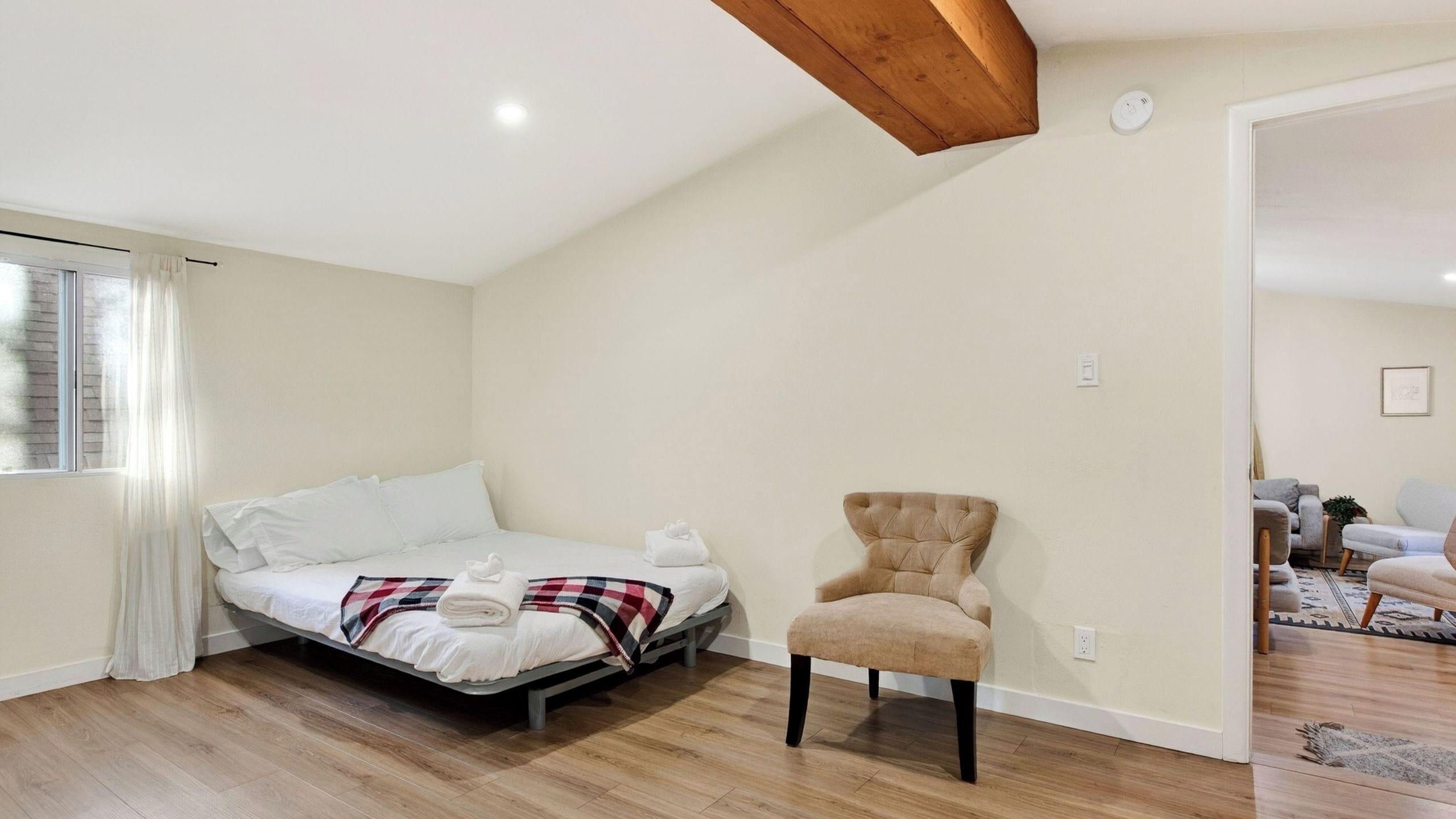 A bedroom with a bed, a chair, a window with curtains, and a wooden ceiling beam.