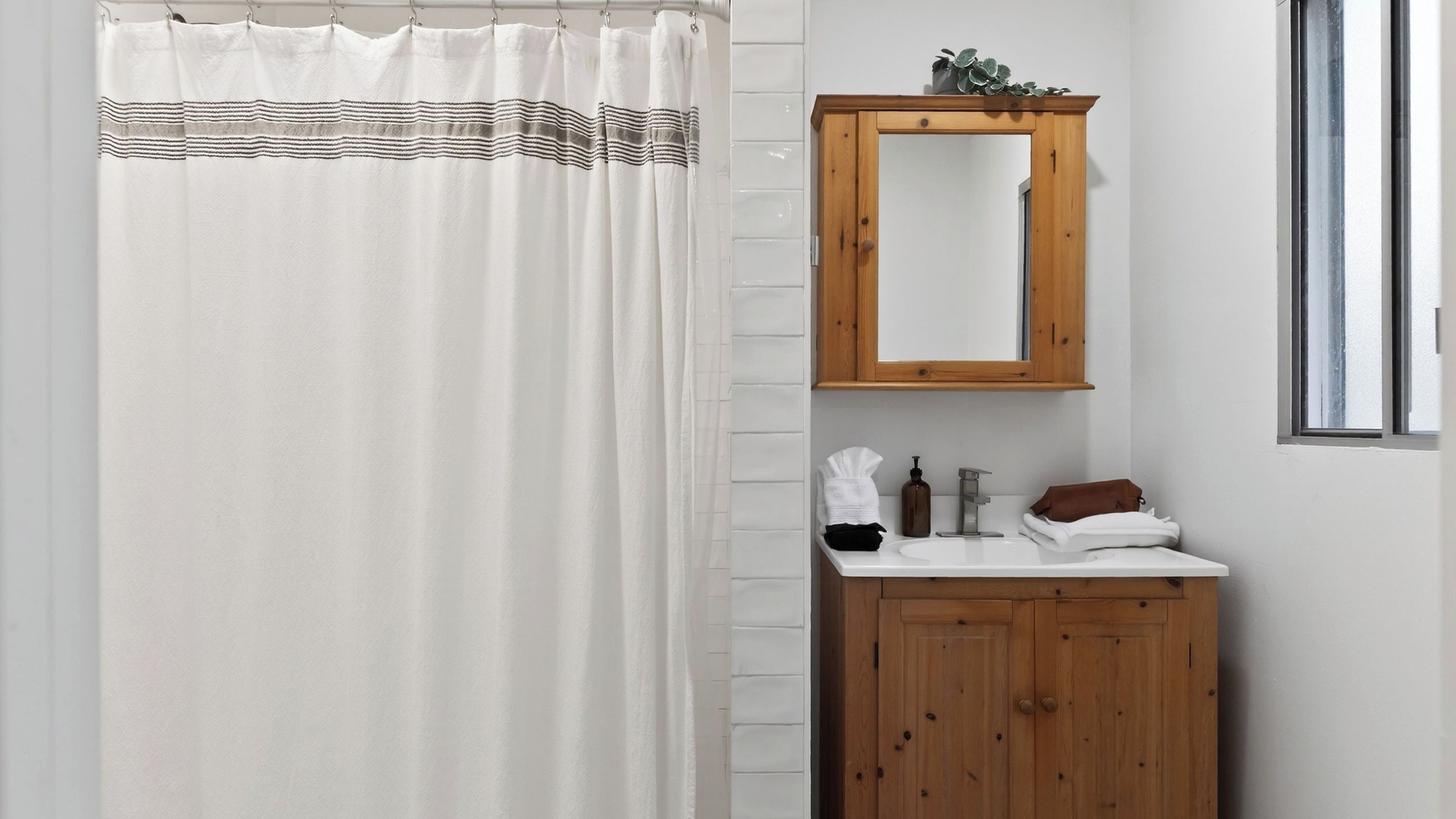 A bathroom with a shower curtain, a wooden cabinet with a mirror, a sink, and a window.