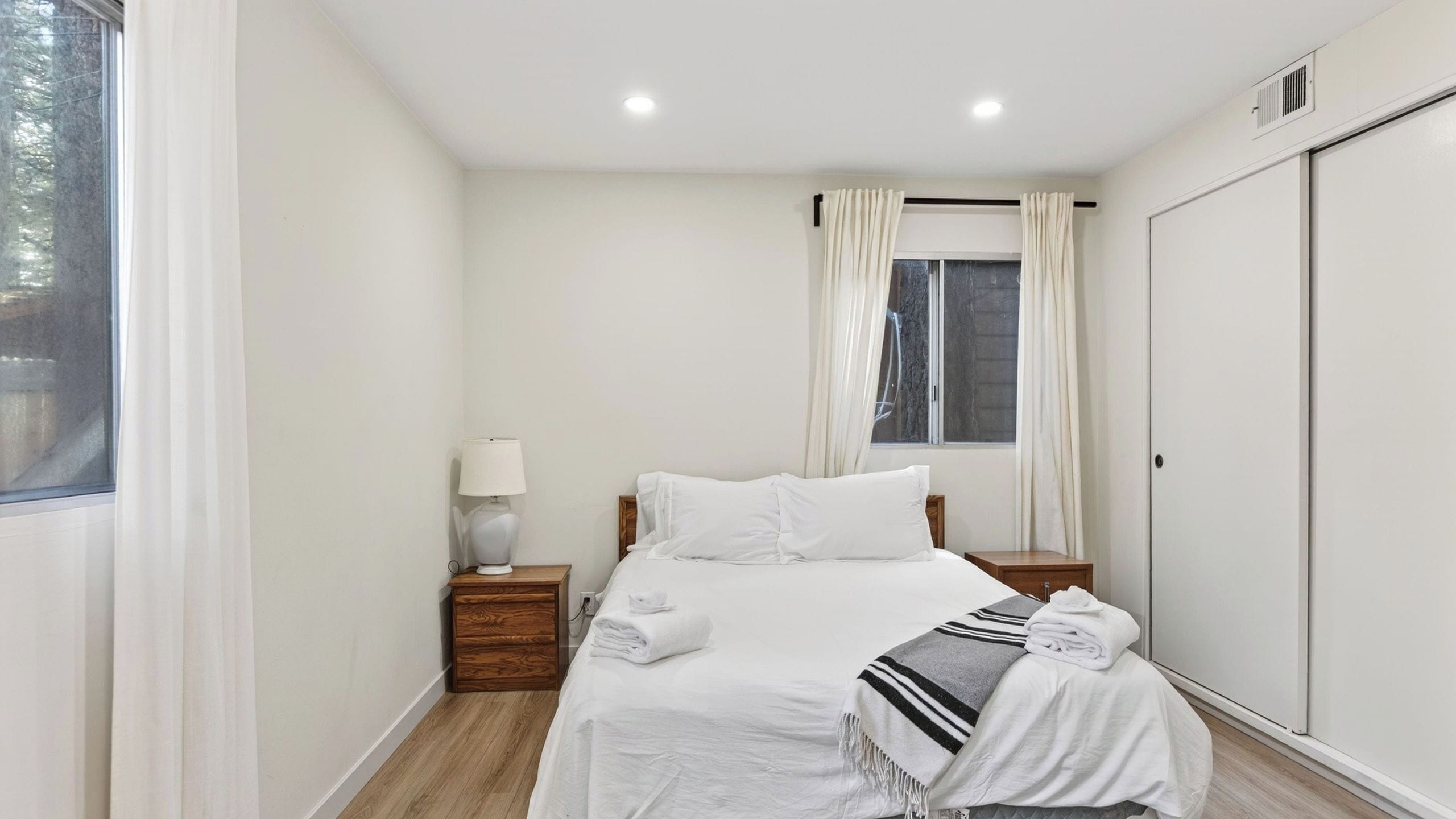 Un dormitorio con cama, mesitas de noche, una lámpara, una ventana con cortinas y un armario.