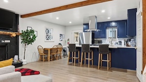 A modern kitchen with blue cabinets, a central island with bar stools, a dining area with wooden chairs, and a fireplace.