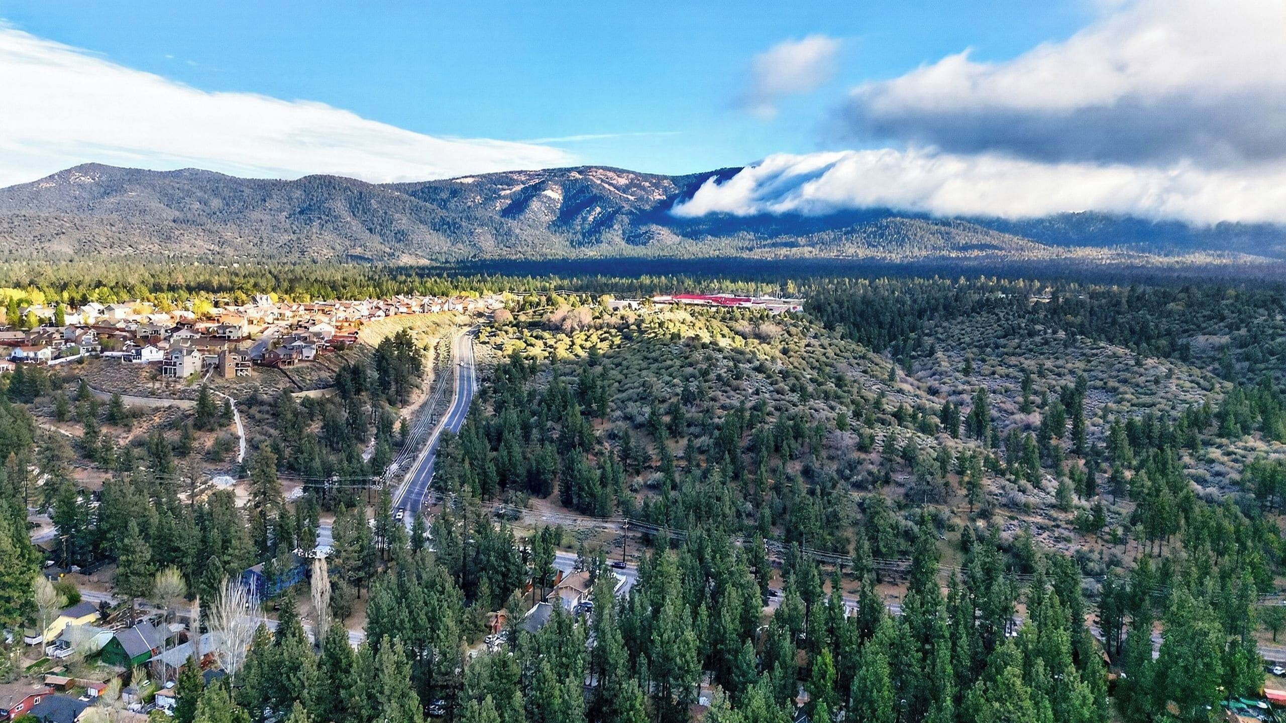 Vue aérienne d’une ville avec une rue principale, une forêt dense et des montagnes en arrière-plan.