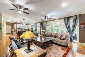 A spacious living room with a sofa, coffee table, and dining area. There are ceiling fans, a kitchen in the background, and large windows with curtains. - 4BR House Near Lake - Hot Tub, Arcade & Fire Pit (Lake Norman of Catawba)