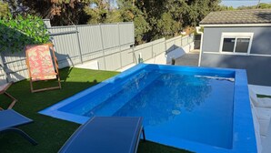 Piscina retangular com interior azul, rodeada por grama sintética, cerca de metal e um pequeno deck com uma cadeira.