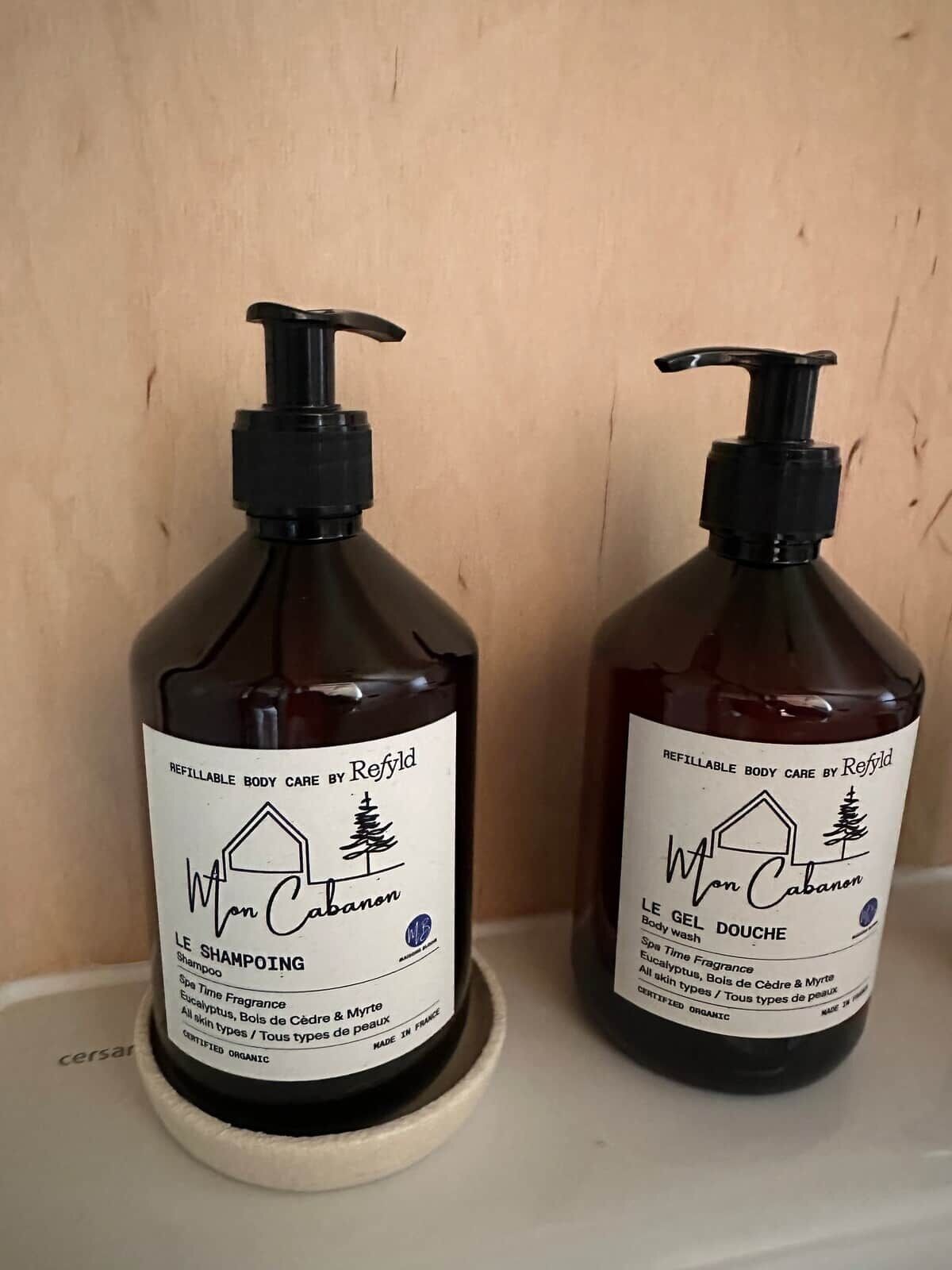 Deux flacons de produits de soins pour le corps rechargeables de la marque Refillable, posés sur une étagère.