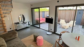 Uma sala de estar moderna com sofá, puf e mesa redonda. Há uma escada, uma televisão e uma porta de vidro deslizante que dá para uma área de estar ao ar livre.