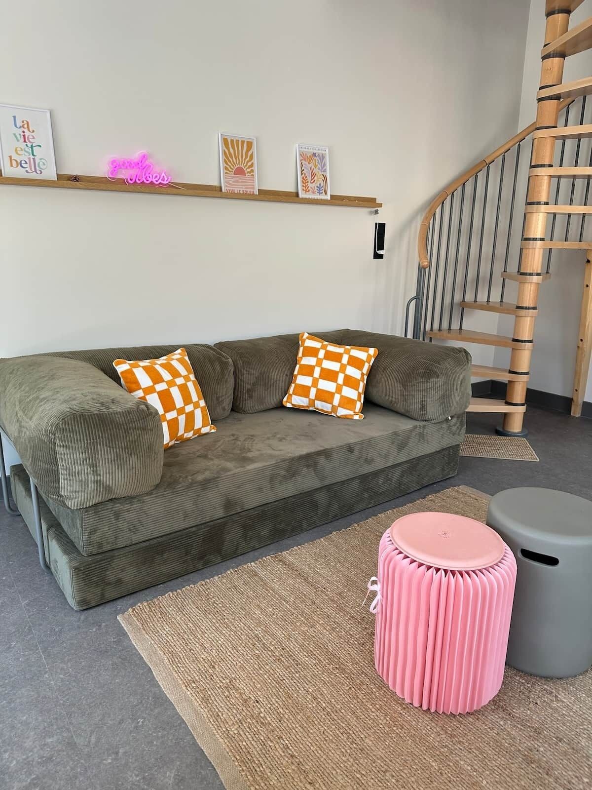 Un salon avec un canapé vert, des coussins à carreaux, un pouf rose, une poubelle grise et un escalier en bois.