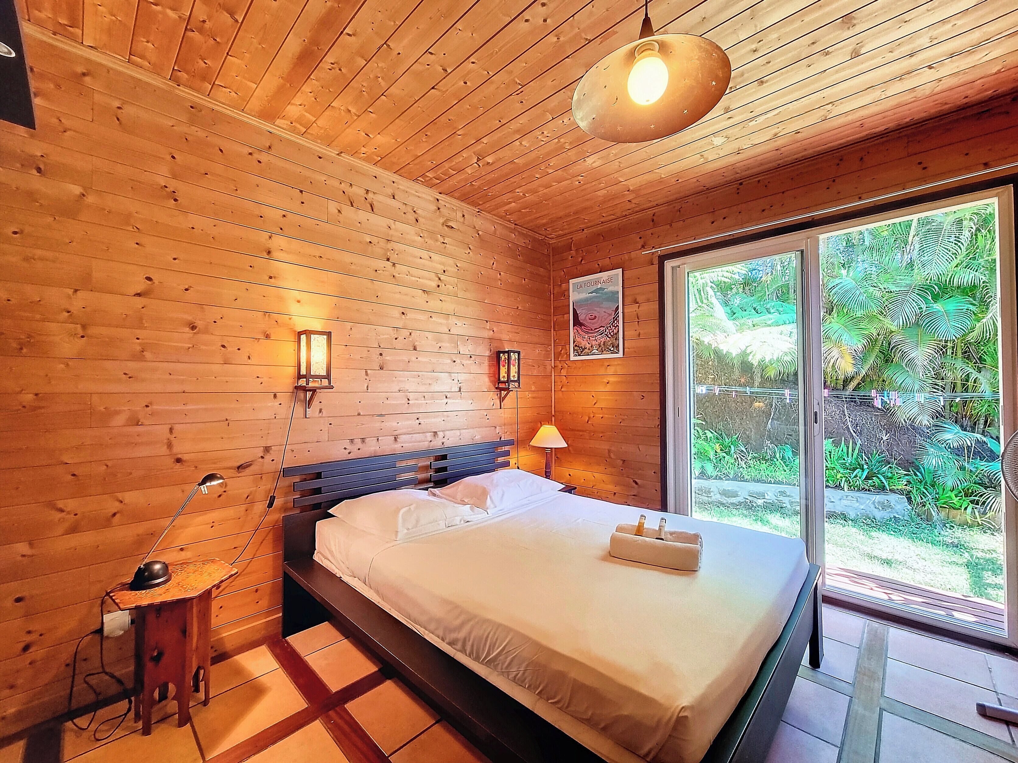 Habitación con paneles de madera, una cama, dos lámparas fijadas a la pared y un ventanal con vistas al verdor.