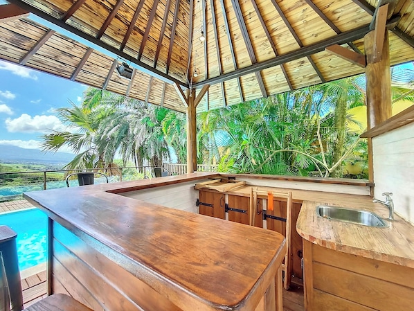 A wooden bar area with a view of a swimming pool and tropical vegetation. - Verger Suspendu - Piscine - Petfriendly (L'Étang-Salé)