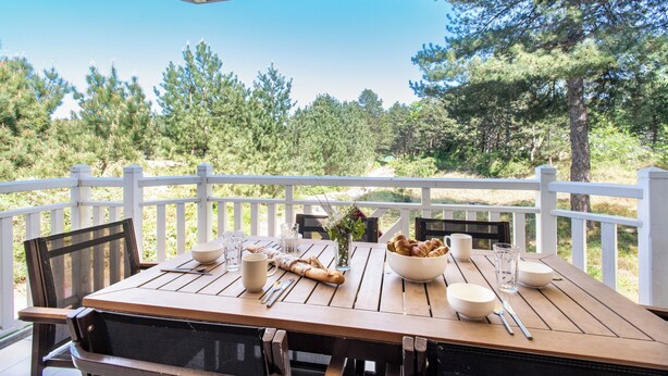 Ett träbord uppdukat utomhus, med ett vitt staket, en skål med bröd och en vas med blommor.