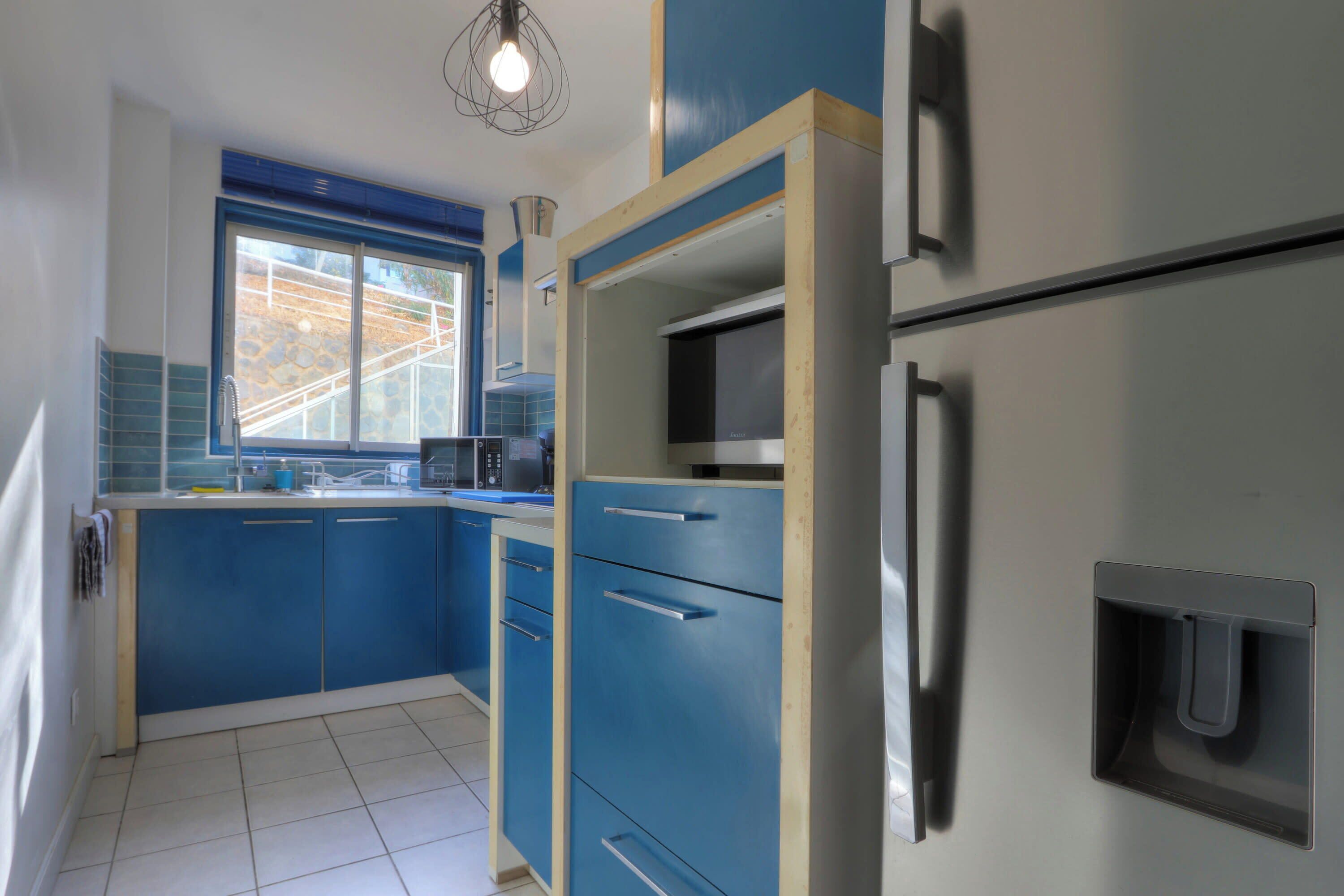 A kitchen with blue cabinets, a stainless steel refrigerator, and a window with a view of a stone wall.