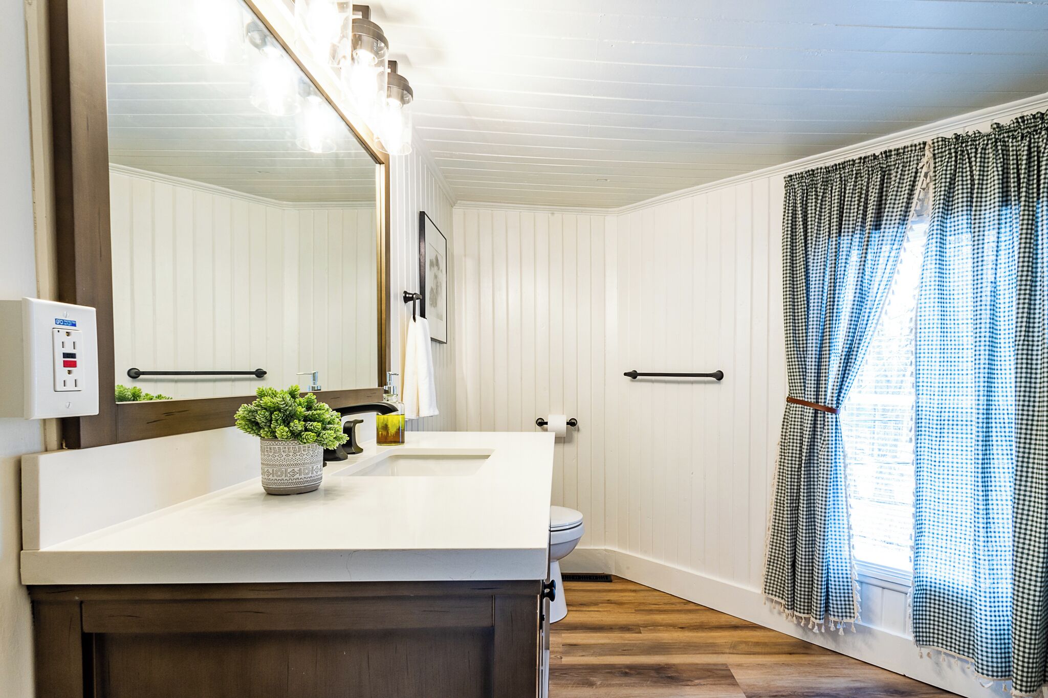 Un baño con tocador blanco, espejo, planta en maceta, inodoro y cortina de ducha.
