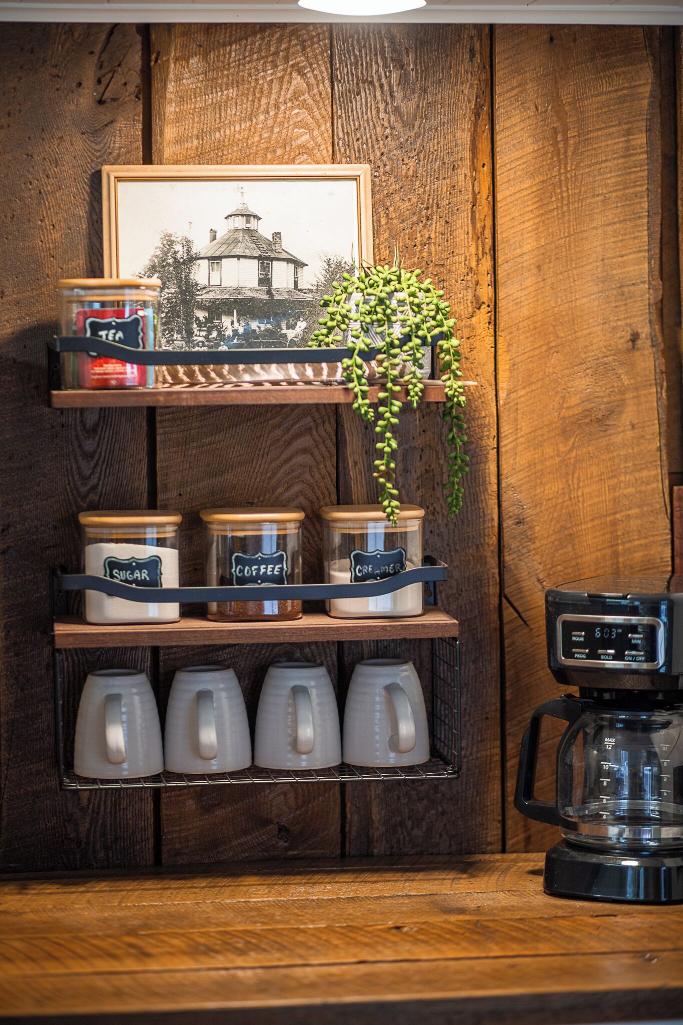 A coffee station with labeled jars of sugar and coffee, a coffee maker, and a framed picture on a wooden wall.
