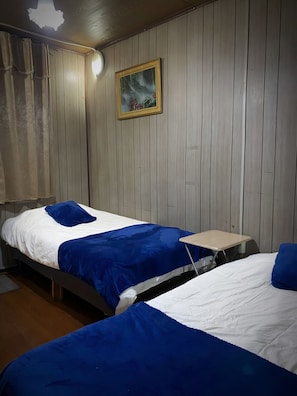 Ein Zimmer mit zwei Betten, jeweils mit einer blauen Decke, einem kleinen Tisch und einem Bild an der Wand.