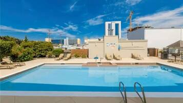 A rooftop swimming pool with a clear blue sky, surrounded by buildings and greenery.