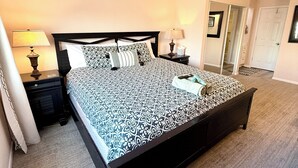 Un dormitorio con una cama grande, dos mesitas de noche, una mesilla auxiliar, una puerta y un pasillo que conduce a otra habitación.