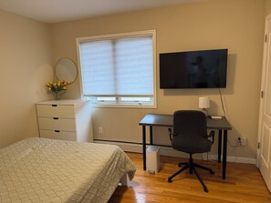 Ein Schlafzimmer mit Bett, Schreibtisch, Stuhl, Fernseher, einem Fenster mit Jalousien, einem Spiegel und einer Vase mit Blumen.