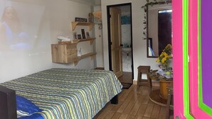 Un dormitorio con una cama, estanterías de madera, una mesita con un florero y una puerta que da a otra habitación.