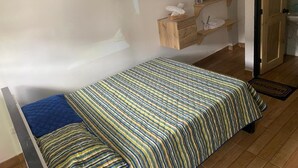 Una cama individual con ropa de cama a rayas, un estante de madera con varios objetos y un tapete de bienvenida.