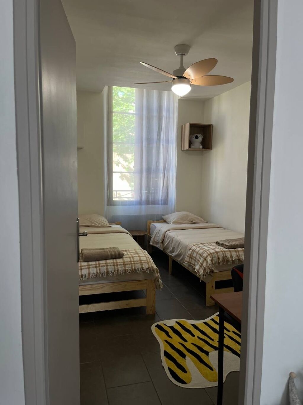 A small, single-room accommodation with two beds, a ceiling fan, a window with sheer curtains, and a zebra-patterned rug.
