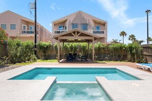 A swimming pool with a covered patio area, a two-story building with balconies, and a fenced yard with greenery. - New remodel w/Pool & feet from the beach! (South Padre Island)