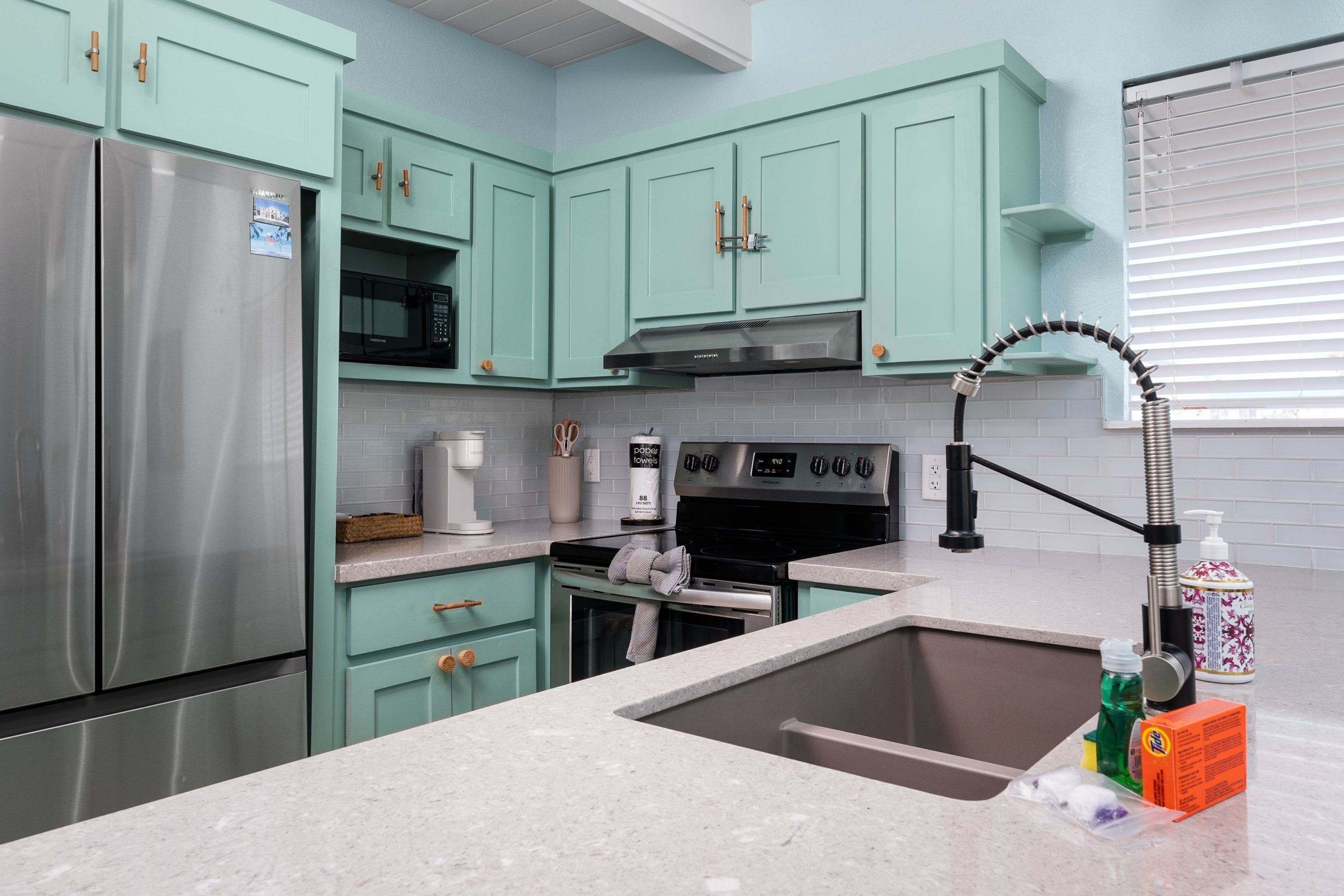A kitchen with teal cabinets, stainless steel appliances, a granite countertop, and a window with blinds.