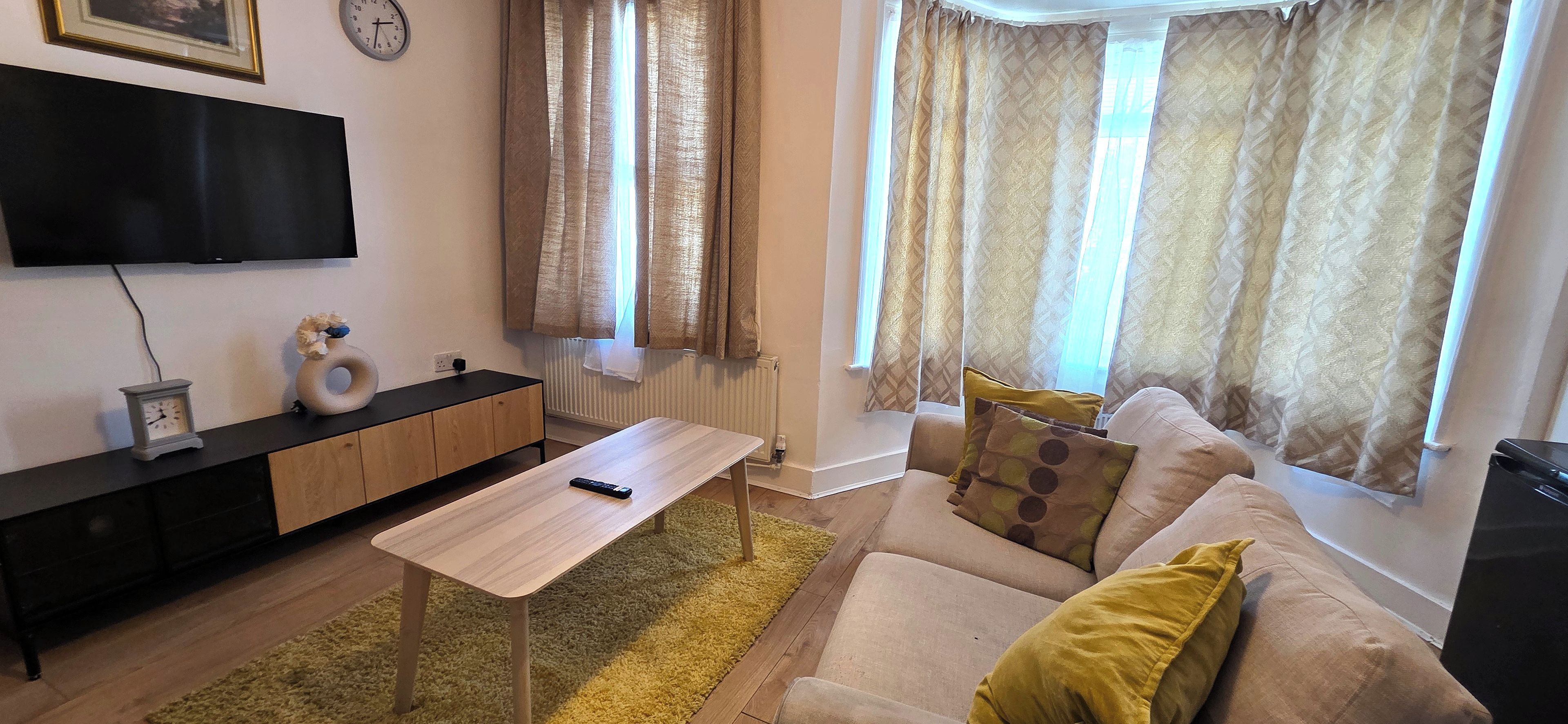 Uma sala de estar com sofá, mesa de centro, TV na parede e janela com cortinas.