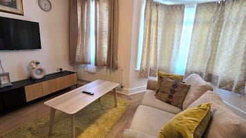 Uma sala de estar com sofá, mesa de centro, TV na parede e janela com cortinas.