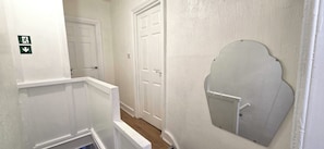 Un pasillo estrecho con paredes y puertas blancas, un espejo pequeño en la pared derecha y una señalización de salida de emergencia verde en la izquierda.