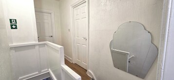 A narrow hallway with white walls and doors, a small mirror on the right wall, and a green emergency exit sign on the left.