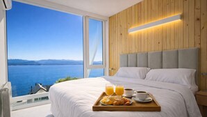 Una camera da letto con un letto grande, una testata in legno, un vassoio con croissant, succo e caffè, e vista su un lago attraverso ampie finestre.