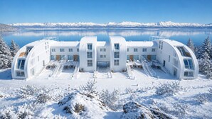 Un edificio moderno, bianco e a più piani, dal design curvo, situato vicino a un lago con montagne innevate sullo sfondo.