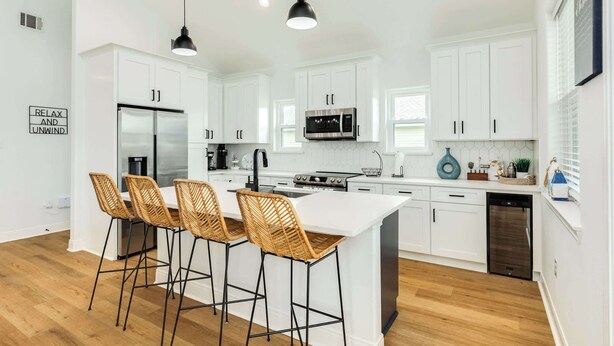 Una cucina moderna con mobili bianchi, un'isola centrale con sgabelli da bar, elettrodomestici in acciaio inossidabile e illuminazione a sospensione.