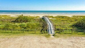 Una spiaggia con una passerella in legno che conduce alla riva, circondata da vegetazione e un cielo sereno.
